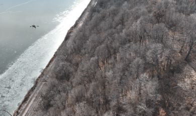 A drone flies above a rail line surrounded by trees and a river.